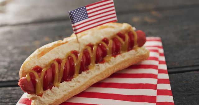 Classic hot dog with a miniature American flag decoration on top, ketchup and mustard in a zigzag pattern, placed on a red and white striped napkin. Ideal for promotions focusing on national holidays like Independence Day, Memorial Day, or any event that promotes American culture and cuisine.