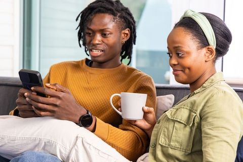 African american couple sharing smartphone and coffee on cozy sofa, relaxing at home
