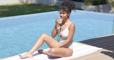 Woman relaxing at pool edge drinking lemon water in striped swimsuit on backyard deck