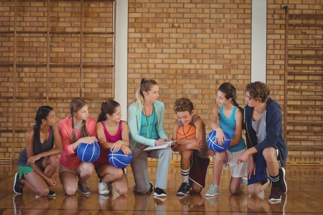 A female coach is seen mentoring a group of high school students holding basketballs in a gymnasium. This image is ideal for use in articles or promotional materials related to youth sports, coaching, physical education programs, or teamwork in sports. It highlights the importance of guidance and support in sports training and the role of a coach in developing young athletes.