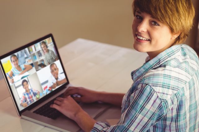 Teenager wearing plaid shirt attending an online class through a video conferencing platform. The screen displays multiple students in a grid layout, each engaged in learning activities from different locations. This image is perfect for articles and resources about online education, virtual classrooms, remote learning, technology in education, and student engagement in digital environments.