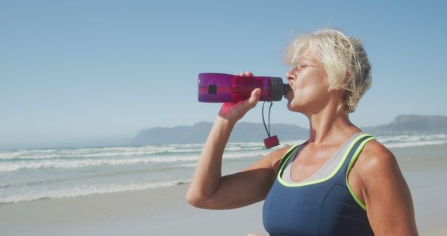 Senior woman drinking water from a sports bottle after exercising on the beach. The ocean waves and clear sky in the background showcase a healthy and active lifestyle. Ideal for use in content related to fitness, hydration, active aging, beach workouts, and health and wellness campaigns.