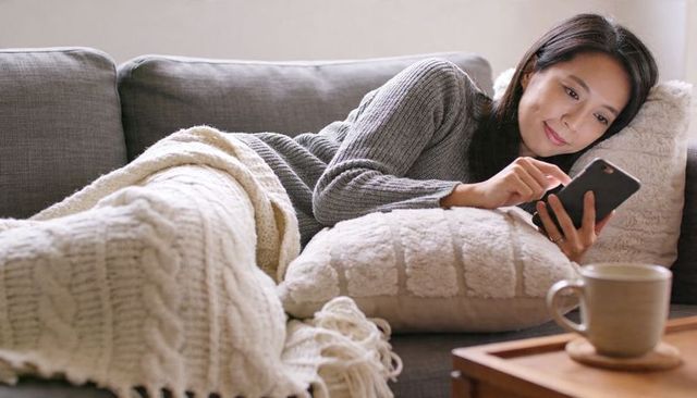 Reclining on sofa scrolling smartphone under chunky knit blanket with coffee mug