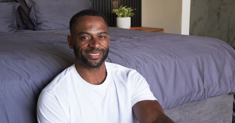 Smiling man relaxing at foot of modern king bed with gray duvet and bedside plant