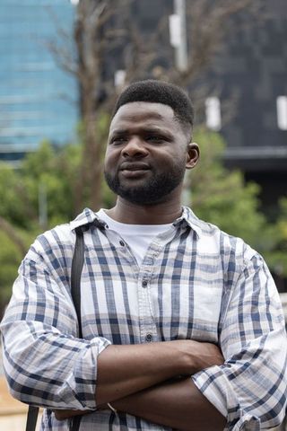 Confident african american man posing with arms crossed wearing plaid shirt and bag in urban plaza