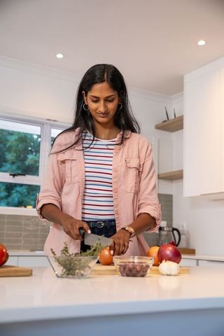 Indian woman chopping onion on kitchen island with chef knife in modern sunlit kitchen
