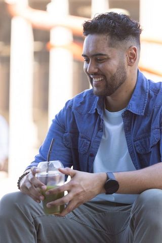 Smiling african american man holding green smoothie on sunny urban cafe patio wearing smartwatch