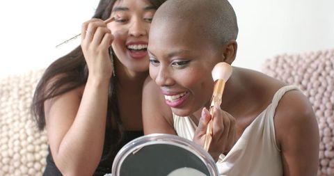Smiling friends applying makeup at home — woman with shaved head using powder brush