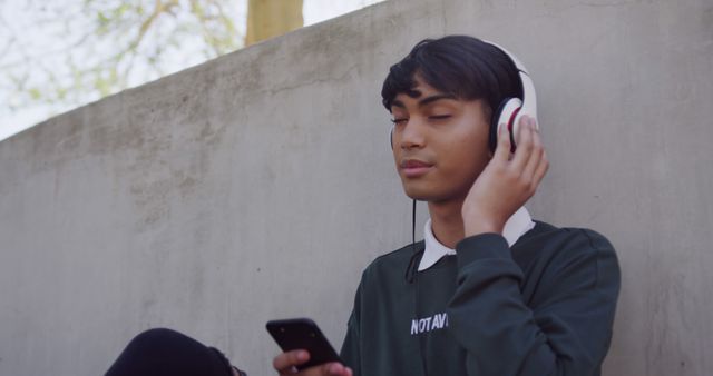 Young man in casual clothing listening to music with headphones while sitting outdoors. Perfect for use in articles on relaxation, music streaming services, technology and lifestyle blogs, and advertisements promoting audio equipment or mobile apps.