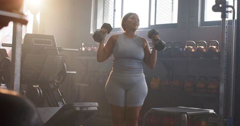 Woman lifting hex dumbbells at shoulder height in sunlit gym near kettlebell rack, strength training