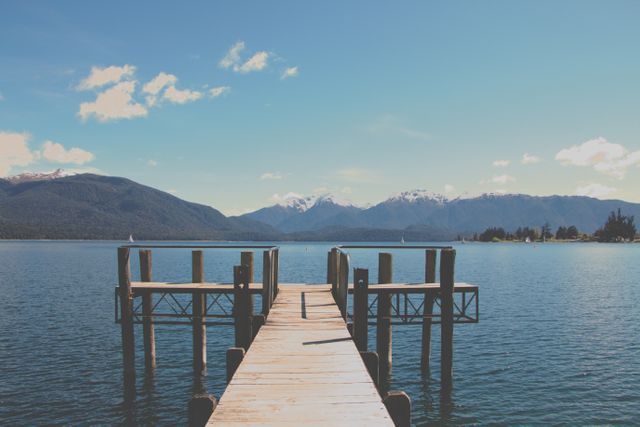 Wooden pier extending over tranquil lake with mountain range in background. Ideal for travel brochures, websites promoting outdoor activities, and relaxation-themed content.