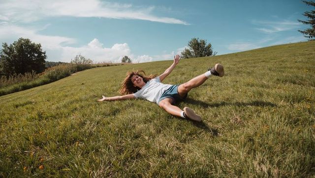 Teenage girl sliding down grassy hill with arms spread on sunny summer day