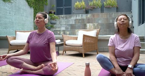 Two women meditating with headphones on patio yoga mats for outdoor mindfulness practice