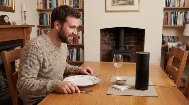 Smiling man sitting at dining table facing smart speaker by fireplace, cozy home automation scene