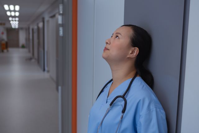 Female doctor in blue scrubs leaning against a wall in a hospital corridor, looking up with a sad expression. This image can be used to depict themes of stress, burnout, and emotional challenges faced by healthcare professionals. Suitable for articles, blogs, and campaigns related to mental health, healthcare industry, and medical staff well-being.