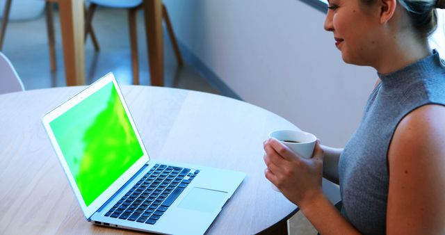 Young woman working on laptop while holding coffee cup in modern office - Download Free Stock Images Pikwizard.com