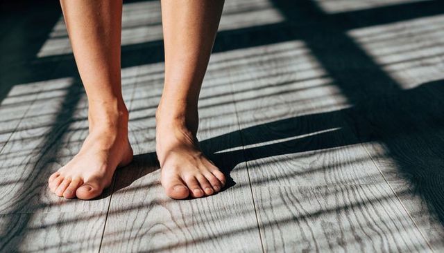 Standing barefoot on sunlit wood floor casting bold slatted blind shadows and warm texture