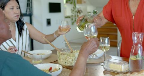 Senior women sharing casual home dinner pouring white wine over pasta salad, dining table