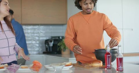 Diverse friends assembling sandwiches at kitchen island, man draining greens with colander