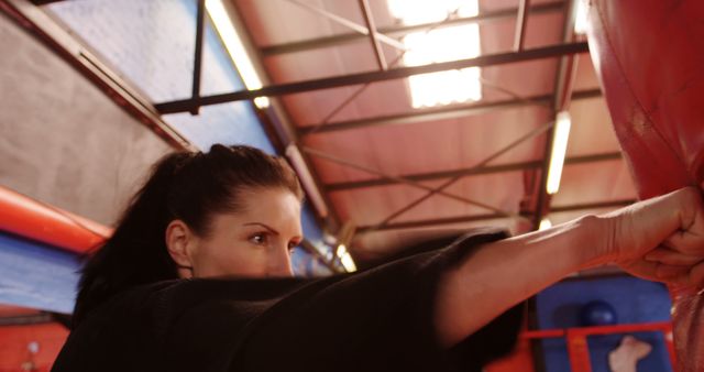 Woman practicing boxing punch in a gym with a red punching bag under industrial ceiling lights. Suitable for content related to fitness, martial arts training, self-defense techniques, female empowerment, athletic inspiration, and health-focused articles.