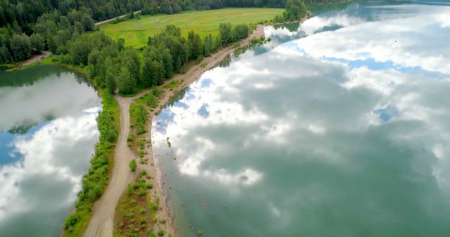 A stunning aerial view capturing a lake with crystal-clear, reflective water mirroring the sky and clouds above. The surrounding lush green forest and an unpaved path run along the lake's edge, adding to the natural beauty and tranquility of the scene. This image is perfect for promoting travel destinations, nature reserves, outdoor activities, and environmental conservation projects.