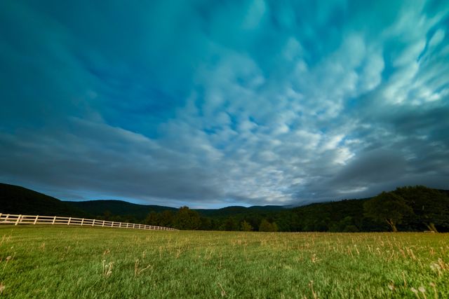 Expansive meadow landscape under a vibrant evening sky with scattered clouds. Rolling hills stretch across the horizon, enhancing the tranquil rural ambiance. Ideal for nature and travel blogs, outdoor magazines, and background settings for presentations promoting peaceful, natural environments.