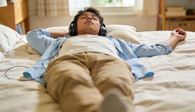 Young man relaxing on bed listening to music with wired over-ear headphones