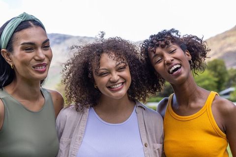 Three diverse women laughing together in sunlit park, enjoying friendship