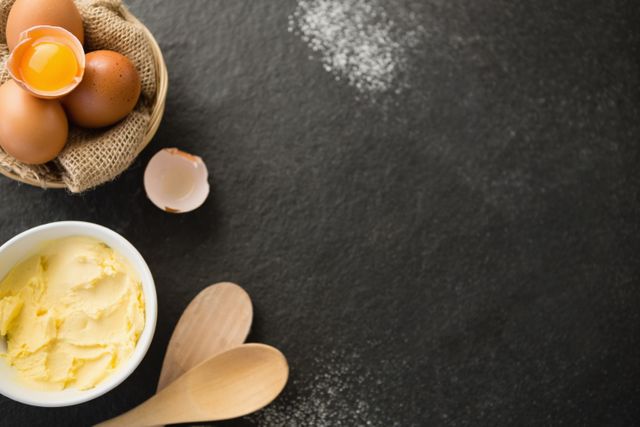 Overhead view showing butter, eggs, and wooden spoons on a dark surface, with a sprinkle of flour around. Ideal for use in cooking or baking tutorials, recipes, food blogs, or culinary-themed websites. Perfect for promoting baking products or kitchen tools.