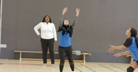 Diverse female volleyball players practicing set and receive drill in school gym with coach