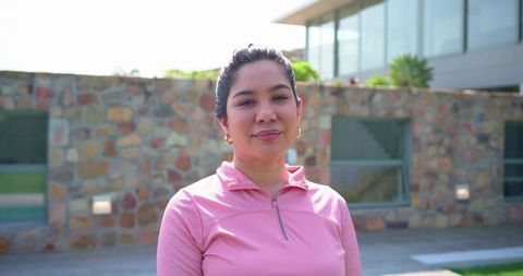 Confident woman standing in sunlit modern courtyard wearing pink activewear and gold hoops
