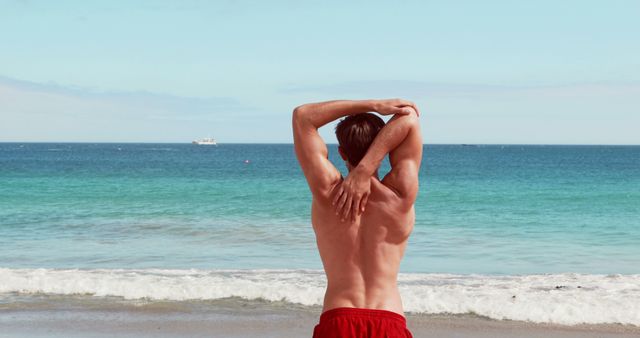 Man stretching with arms above head facing ocean, with calm waves and ship on the horizon. Ideal for health and wellness, fitness, beach activities, summer vacations. Perfect for blog posts, articles, or advertisements focused on fitness, relaxation, and outdoor activities.