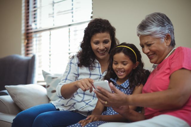 Three generations of women enjoying time together in living room, using mobile phone. Perfect for themes of family bonding, technology in everyday life, and multigenerational relationships.