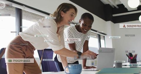 Two businesswomen collaborating over laptop with floating data overlays in modern office