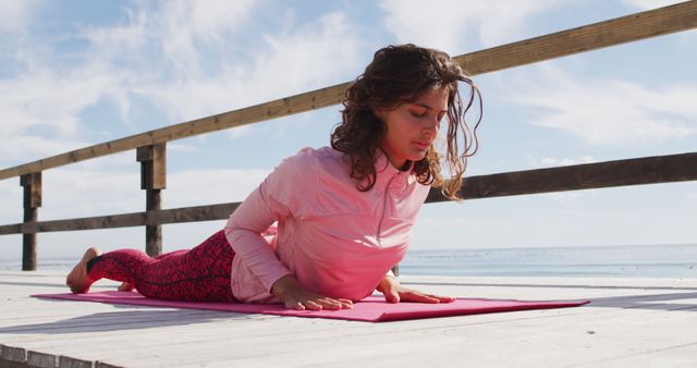 Young woman practicing yoga on a wooden deck by the sea. She is focusing on her breathing and performing a backbend pose on a pink yoga mat. This image is perfect for use in health and wellness campaigns, promoting outdoor activities, or illustrating yoga techniques.