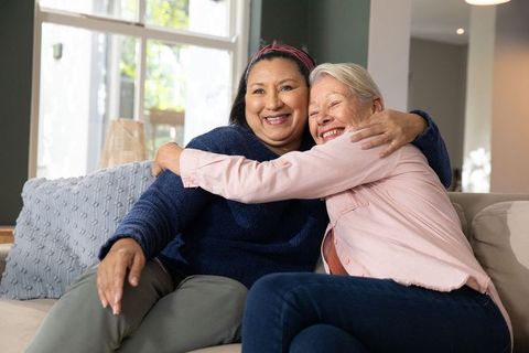 Diverse mature women hugging and smiling on cozy living room sofa, friendship and warmth