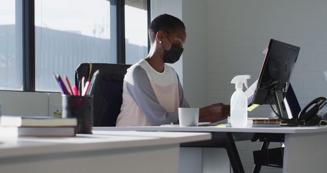 African american businesswoman wearing face mask, sitting at desk at work. independent creative business at a modern office during coronavirus covid 19 pandemic.