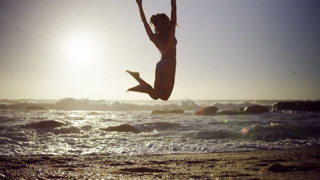 Woman joyfully jumping on sandy beach during golden sunset with ocean waves in background. Perfect for travel promotion, summer vacation advertisements, beach lifestyle blogs, or social media campaign focusing on outdoor fun and freedom.