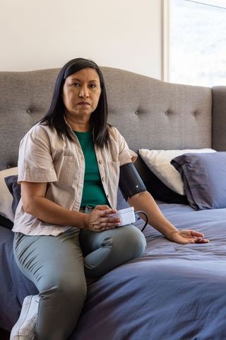 Mature asian woman sitting on bed edge checking blood pressure with portable monitor