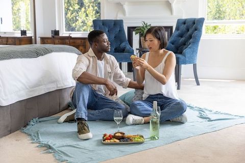 Diverse couple sitting on bedroom blanket sharing smartphone and sipping white wine