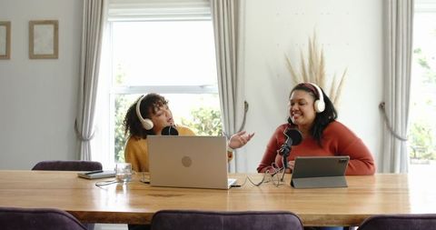 Two women recording casual home podcast with headphones laptop microphones and tablet