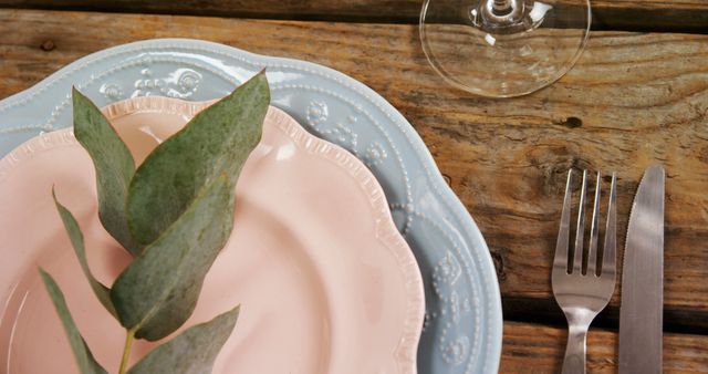 A rustic table setting features a pastel pink plate with a green leaf on it, accompanied by silverware and a wine glass, with copy space. The arrangement suggests a natural and simplistic dining aesthetic, for an outdoor or eco-friendly event.