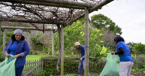 Multicultural volunteers collecting garden debris under wooden pergola, community cleanup