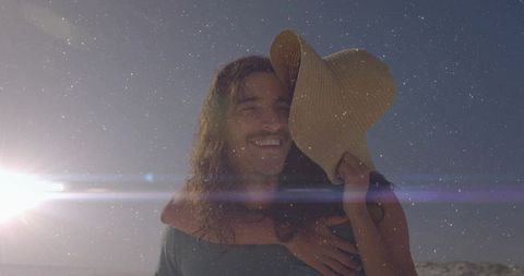 Romantic couple sharing piggyback ride on sunlit beach with sunhat, backlit lens flare