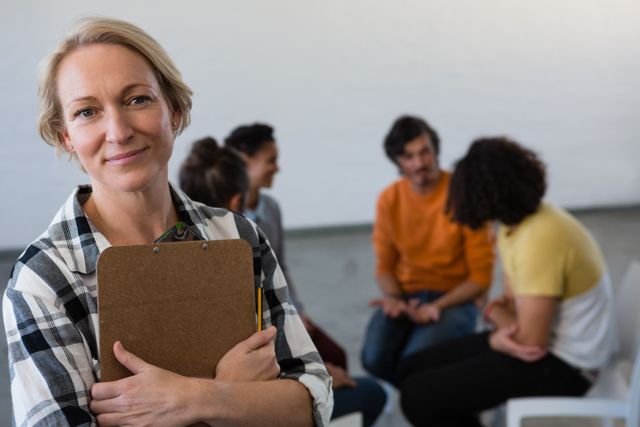 This image depicts a confident teacher holding a clipboard with students engaged in a group discussion in the background. Ideal for use in educational materials, websites, or promotional content highlighting adult education, mentoring, and classroom environments.
