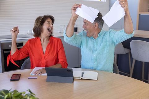 Mature couple celebrating debt-free victory at kitchen table holding torn documents