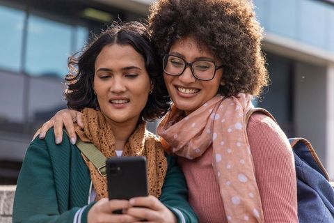 Two young women checking smartphone and smiling on urban campus wearing scarves and glasses