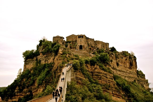 This stock photo showcases an ancient, historic village perched atop a cliff with a dramatic landscape. The stone buildings exude a rustic and medieval charm. A narrow bridge extends from the bottom to the top, where tourists are seen making their way to the village. Ideal for use in travel blogs, historical documentaries, tourism advertisements, and architectural content.