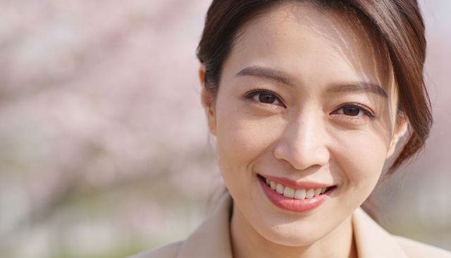 Smiling asian woman wearing beige blazer posing amid spring cherry blossoms