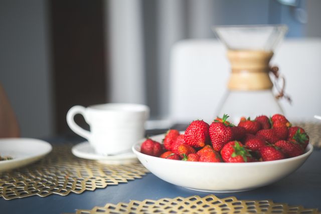 Fresh strawberries filled to the brim in a white bowl, placed on a dining table. Next to the bowl is a white coffee cup and a carafe in the background. Ideal for promoting healthy eating, breakfast recipes, or casual dining settings.